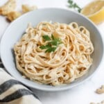 Bowl of Creamy Cauliflower Alfredo pasta in a blue bowl on a white marble benchtop, surrounded by a linen napkin, half lemon and raw cashews.
