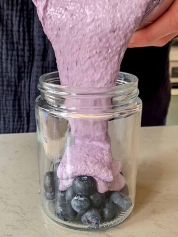 Blueberry blended oats being poured into a jar filled with fresh blueberries.