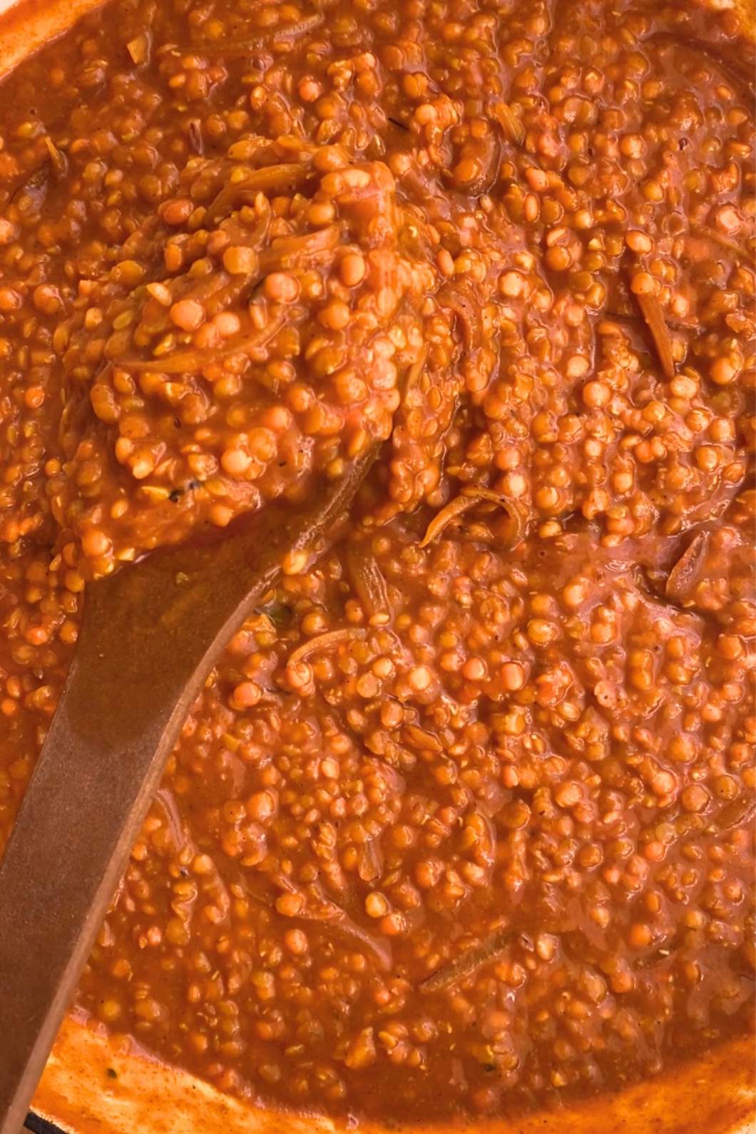 Close-up of a creamy red lentil stew in a pot, featuring a rich tomato base and a wooden spoon partially submerged.