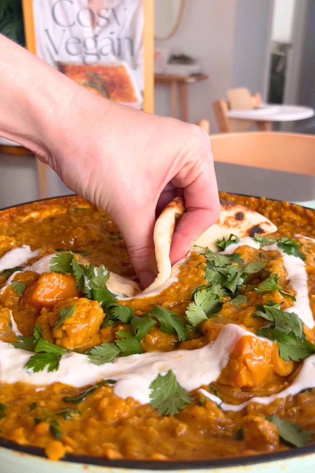 A hand dips naan into a vibrant curry topped with fresh cilantro and yogurt. A "Cosy Vegan" cookbook is blurred in the background.