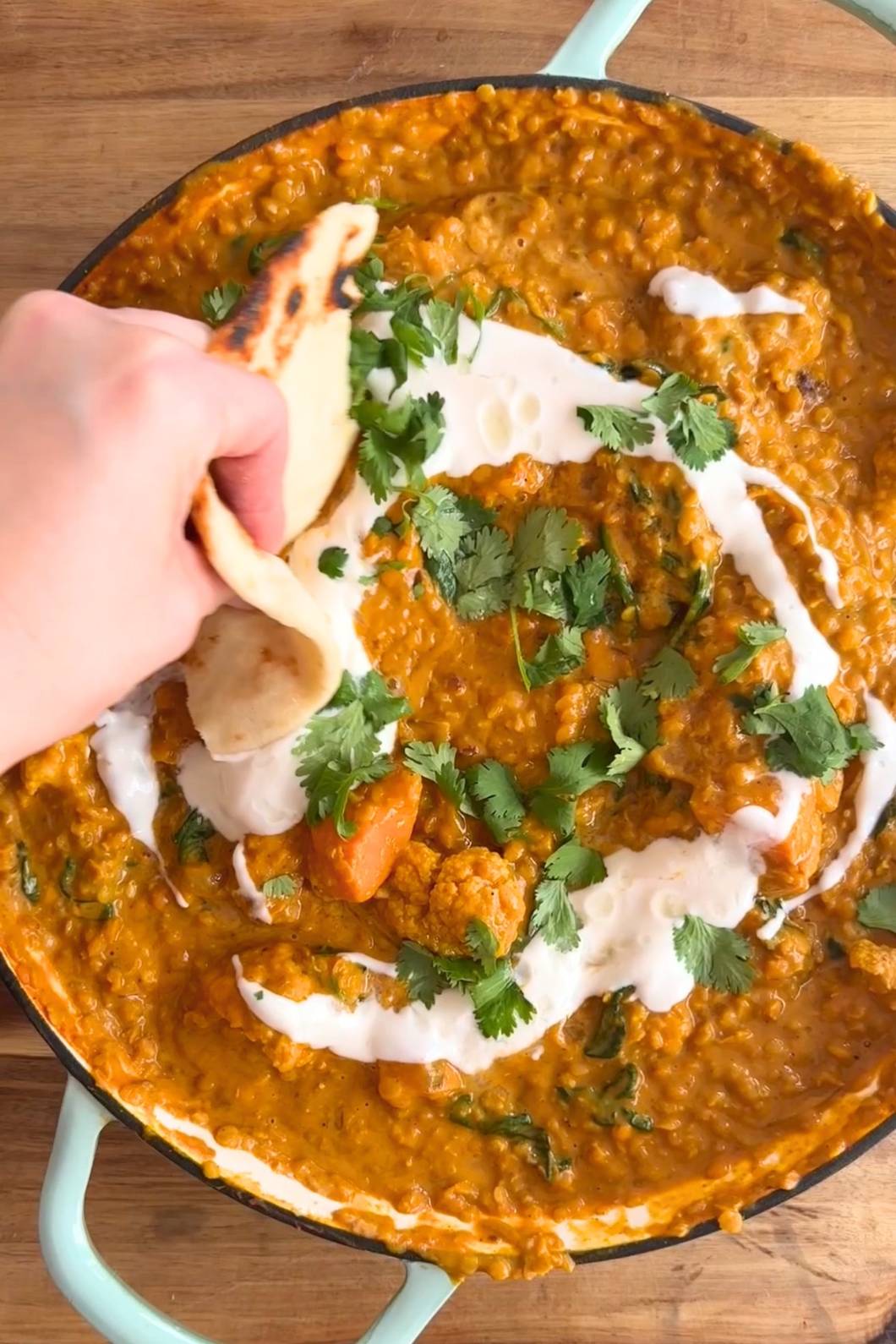 A hand dips naan into a vibrant curry adorned with swirls of cream and fresh cilantro in a skillet.