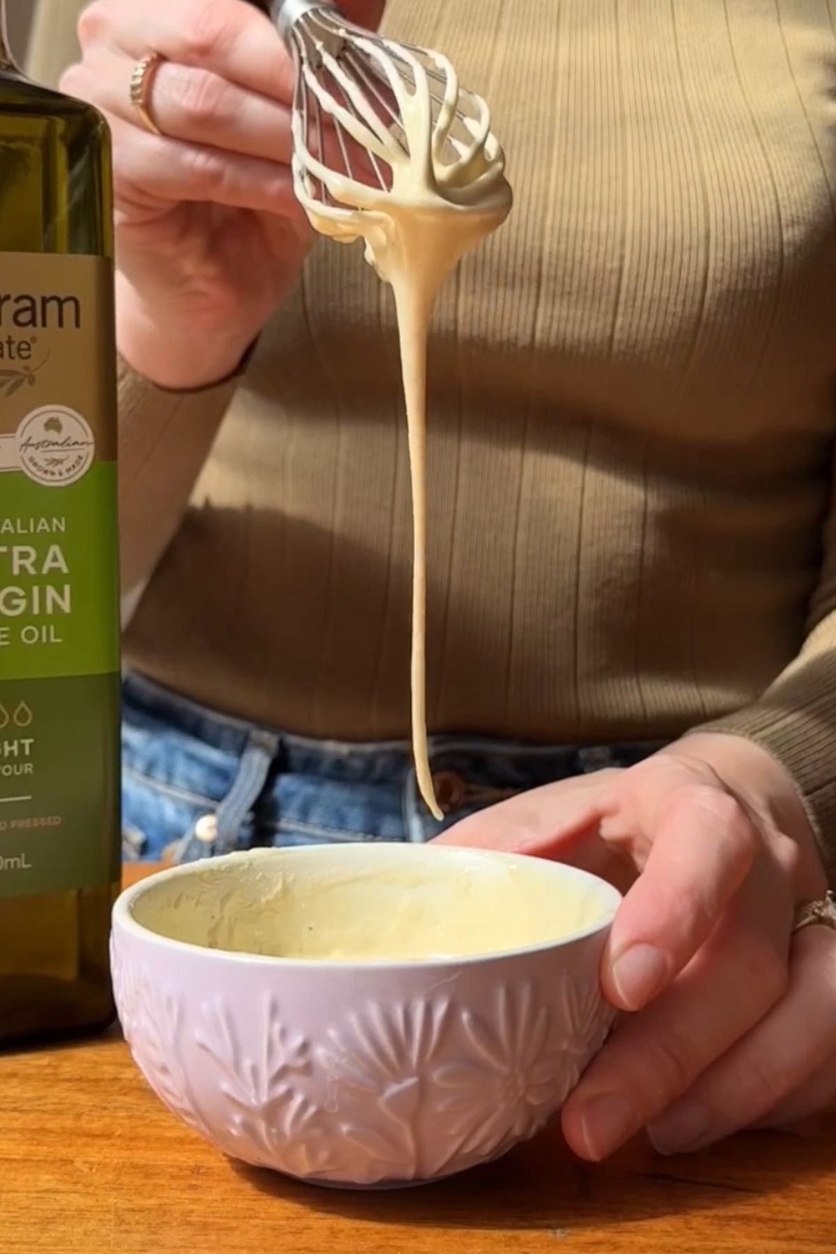 A whisk lifts creamy dressing from a white bowl, with strands dripping back down smoothly.