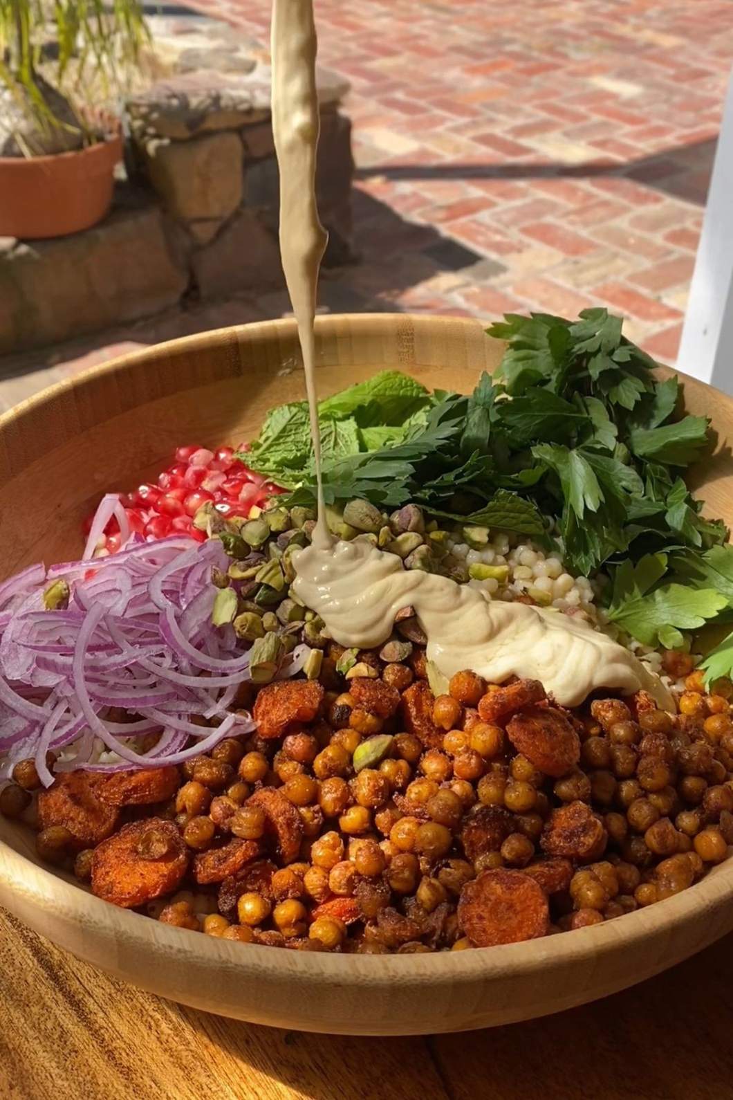 Creamy dressing is being poured over a salad bowl filled with couscous, roasted vegetables, and fresh herbs.