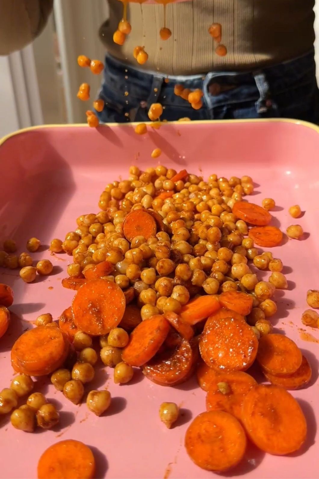 Roasted carrots and chickpeas tumble from a pink tray, coated in a glossy orange seasoning.