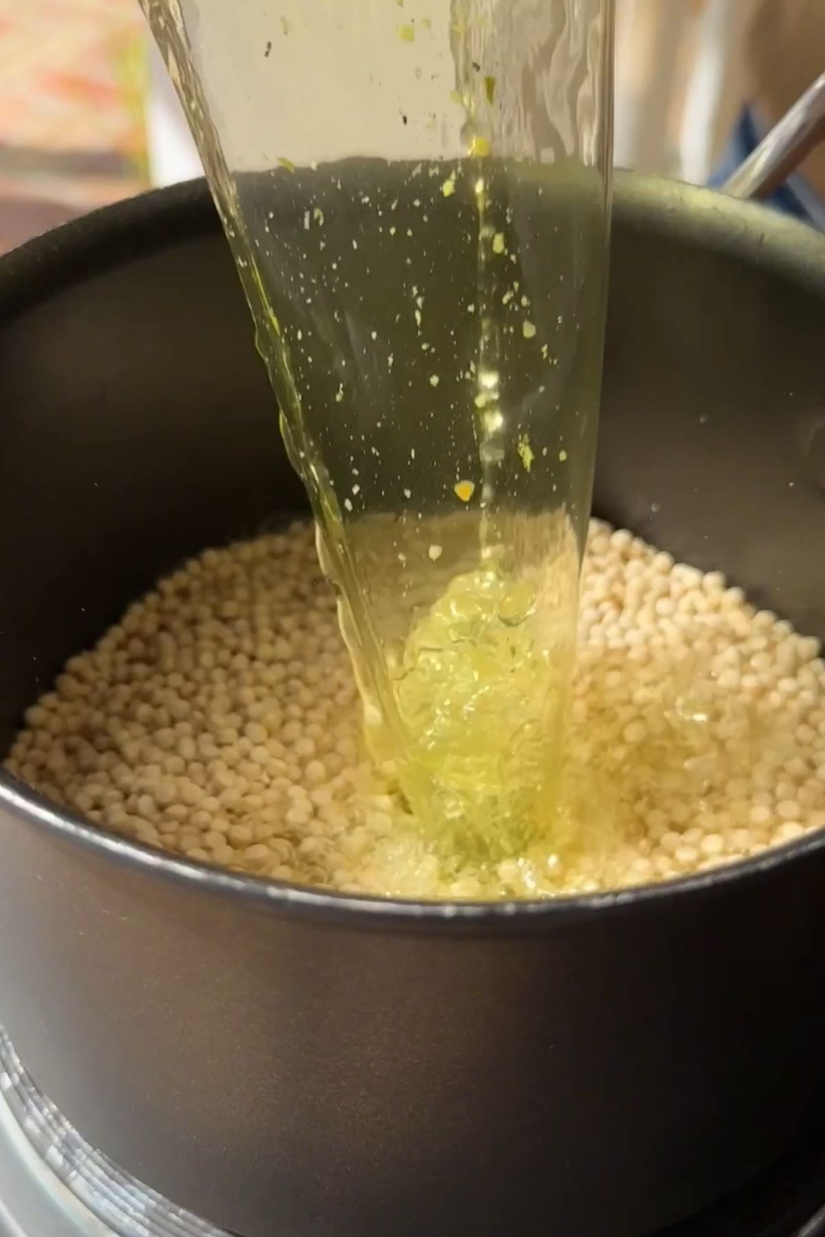 A stream of seasoned broth is poured over dry pearl couscous in a black saucepan.