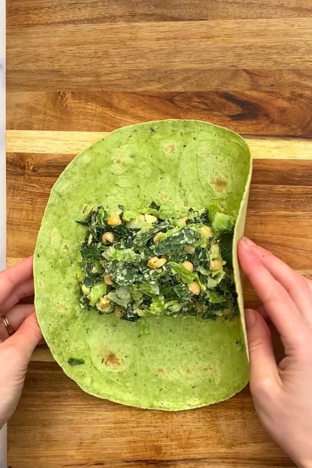 Hands folding a green spinach wrap filled with chickpeas, leafy greens, and creamy dressing on a wooden cutting board.