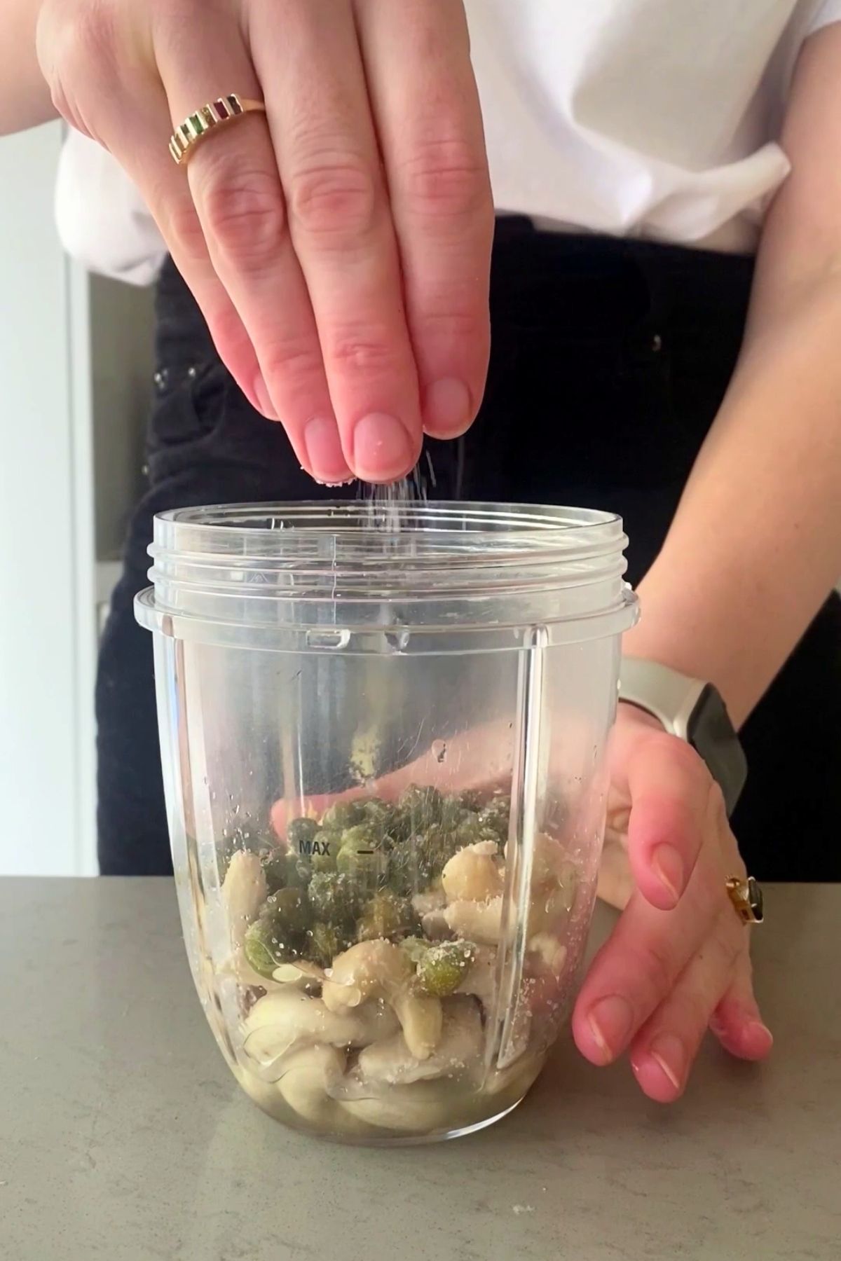 A hand sprinkles salt into a blender cup filled with nuts and greens.