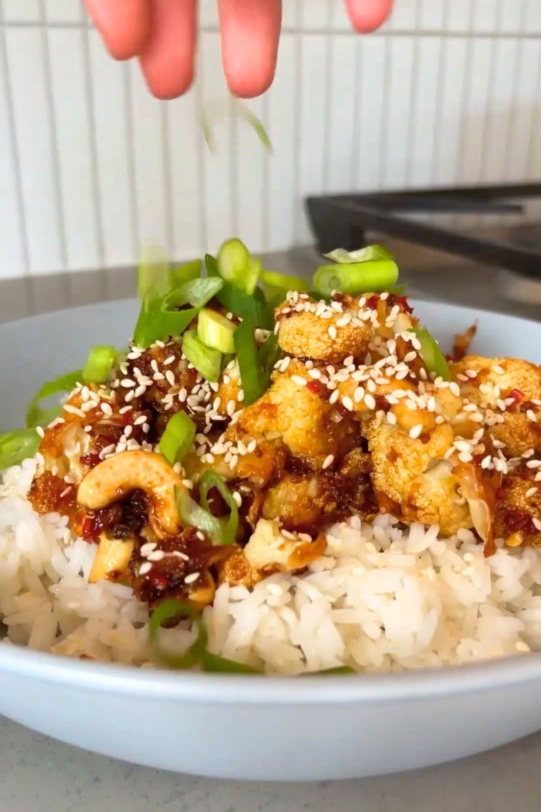 A hand sprinkles sliced green onions over a bowl of rice topped with golden cauliflower and sesame seeds.