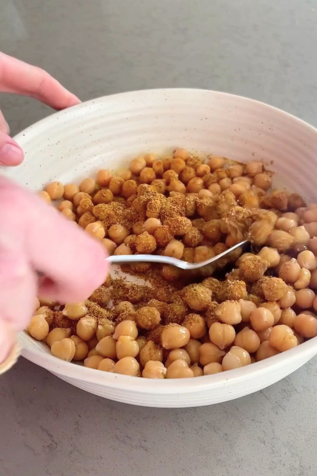 Hands mixing chickpeas and spices in a white bowl with a spoon. 