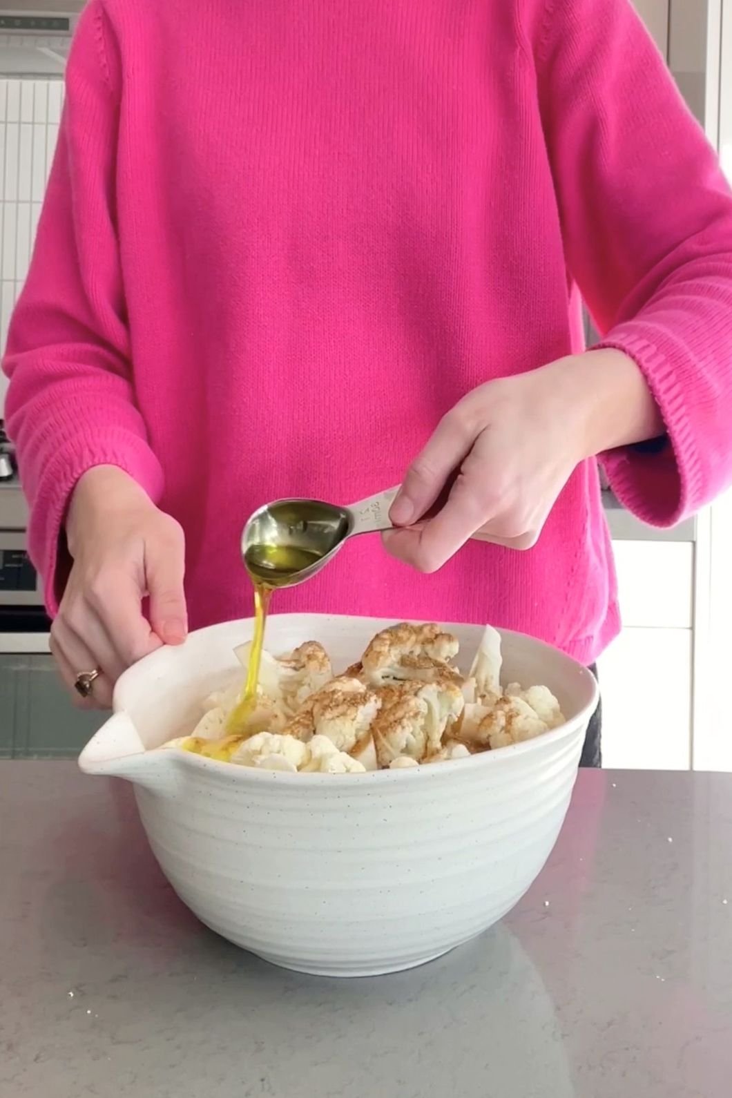 Woman (Liz Douglas) in pink sweater pours olive oil from a tablespoon over cauliflower in a white bowl.