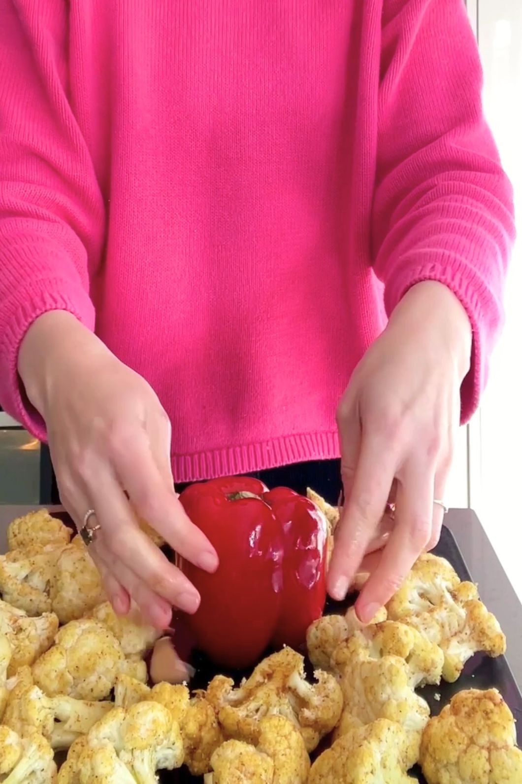 Woman (Liz Douglas) in a pink sweater arranging seasoned cauliflower and a red bell pepper on a baking tray.