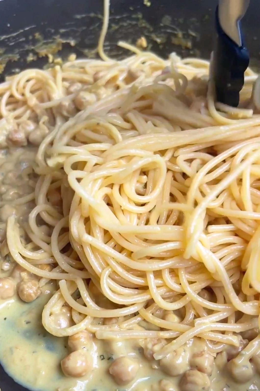 Cooked spaghetti being stirred into a creamy chickpea sauce with tongs.