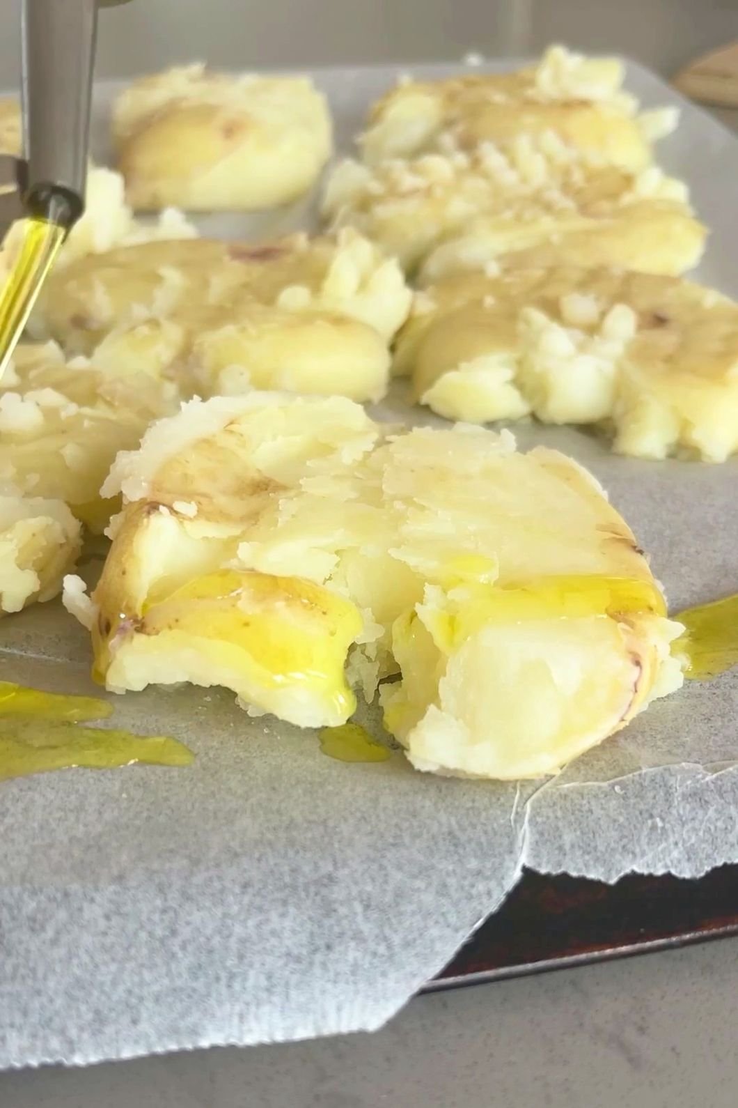 Olive oil being drizzled over flattened boiled potatoes on a tray.