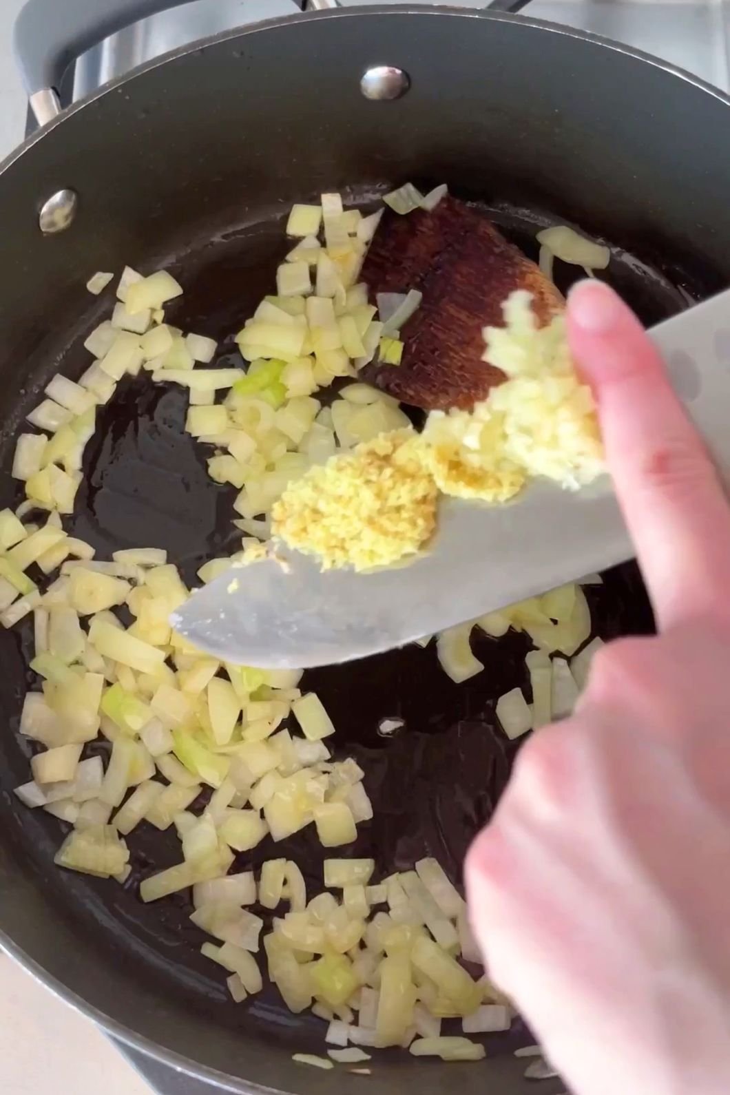 A hand adds minced garlic and ginger to a pan with sautéing onions.