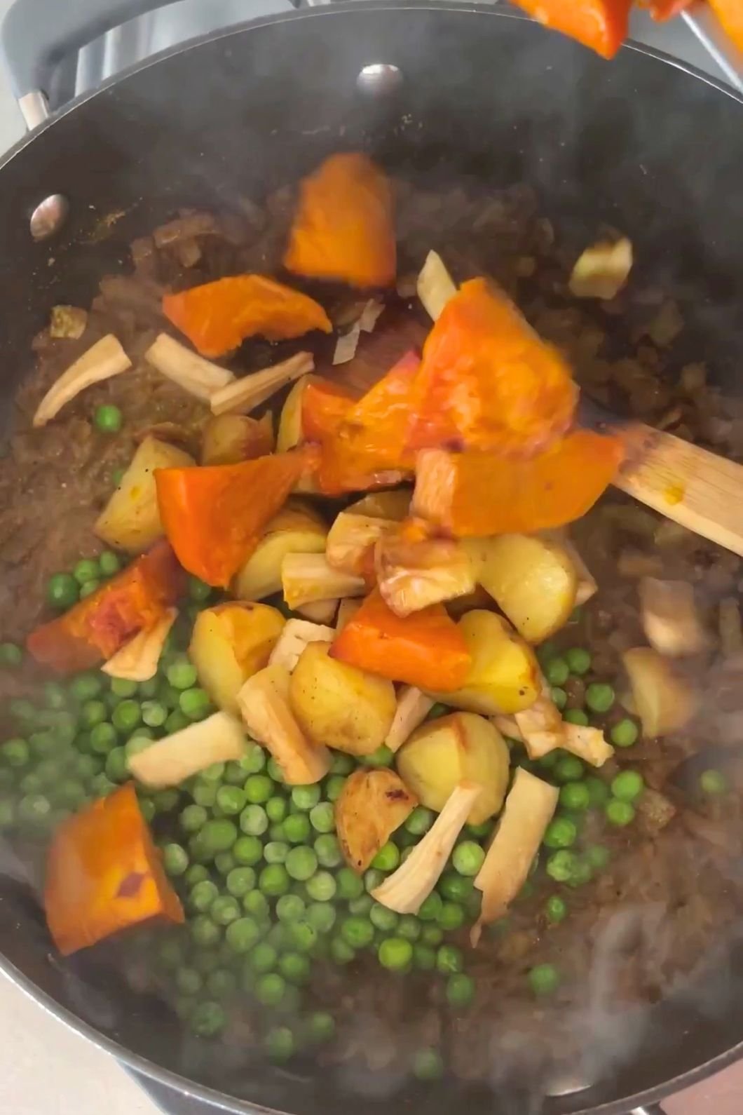 Steaming chopped vegetables and peas are stirred together in a large black pot.
