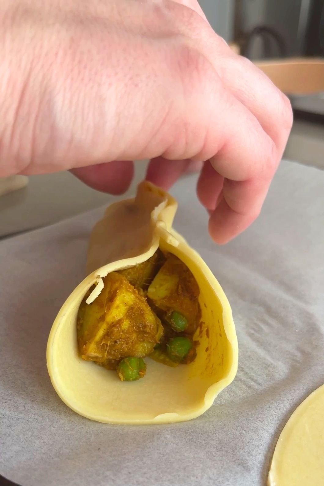 A hand folds pastry around a curried vegetable filling on baking paper.