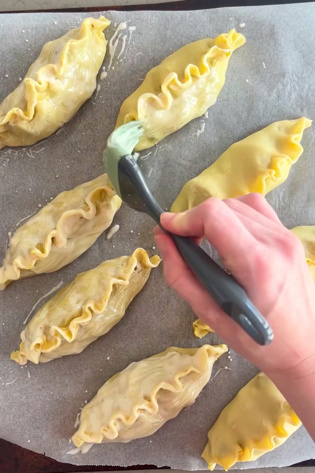 A hand brushes glaze over sealed pastry parcels on a lined baking tray.