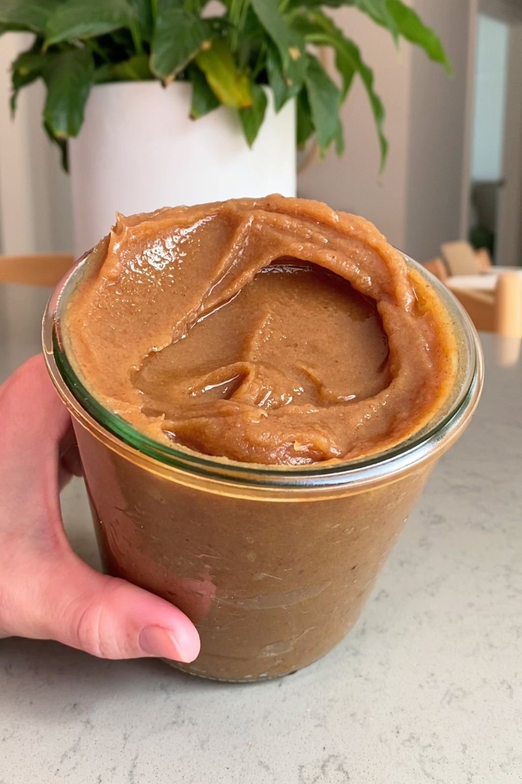 A hand holds a glass jar filled with thick, glossy caramel-colored spread, swirled smoothly on top.