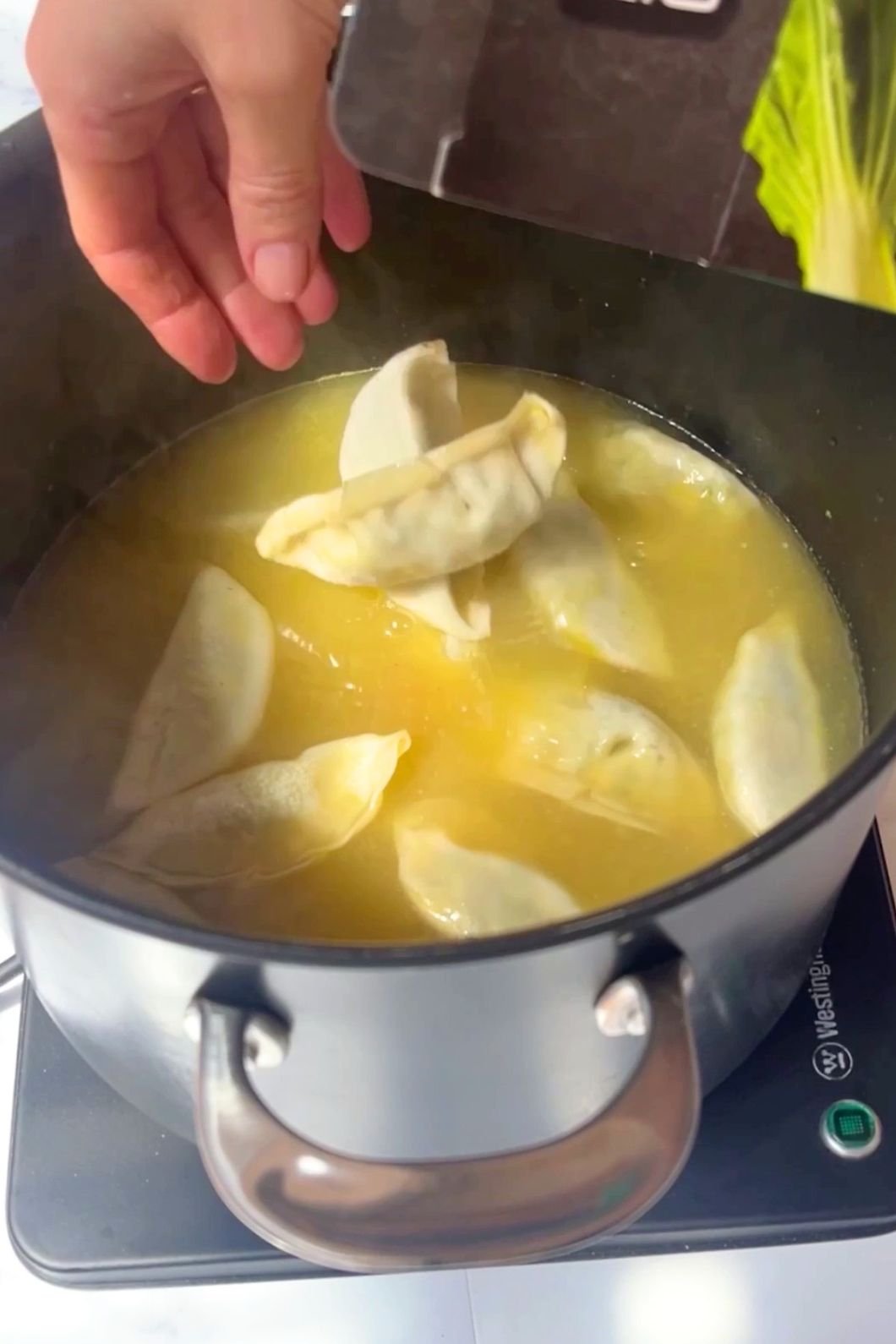 A hand adds dumplings to a pot of yellow simmering broth.