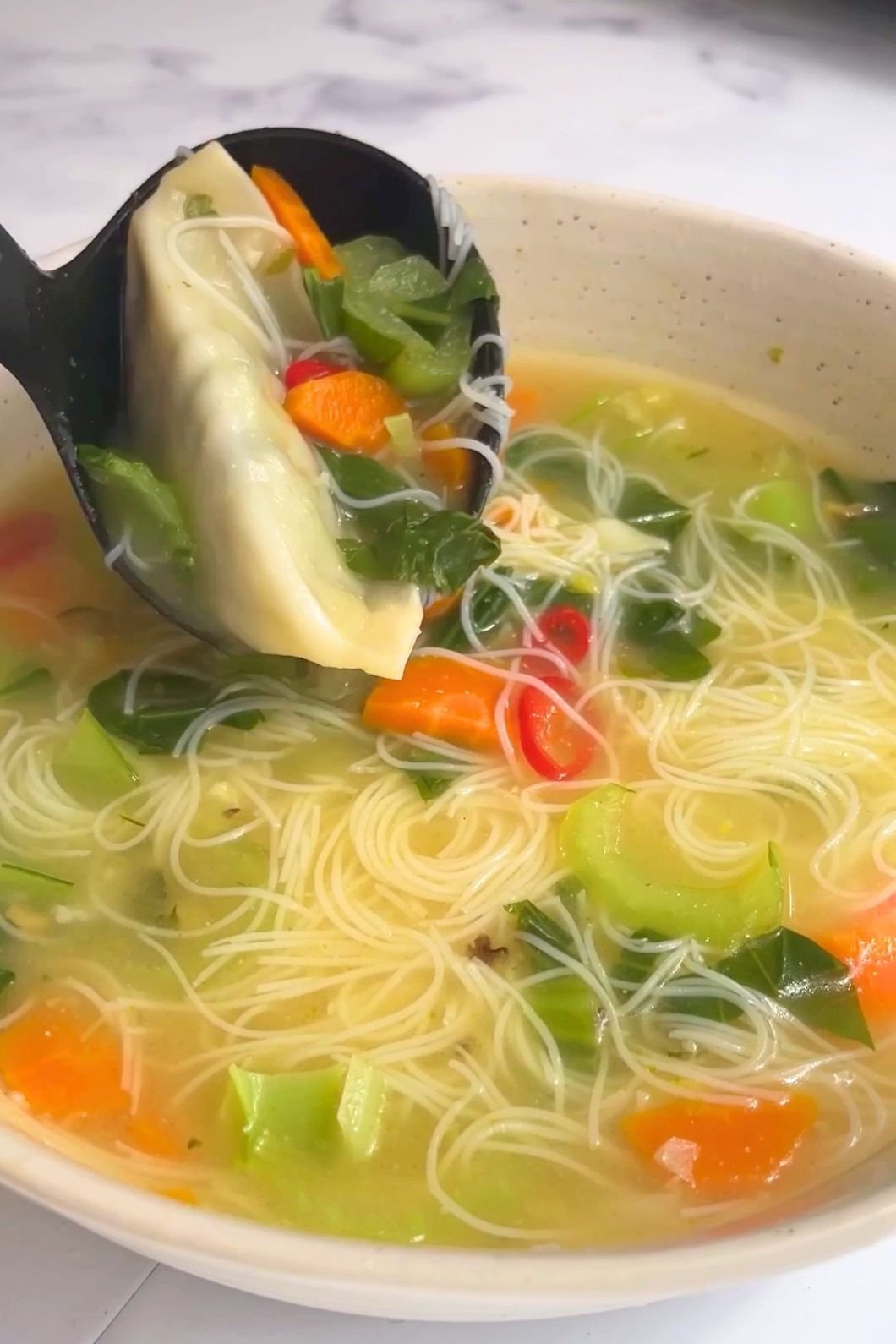 A ladle lifts a dumpling from a bowl of noodle soup with greens, carrots, and red chili slices.