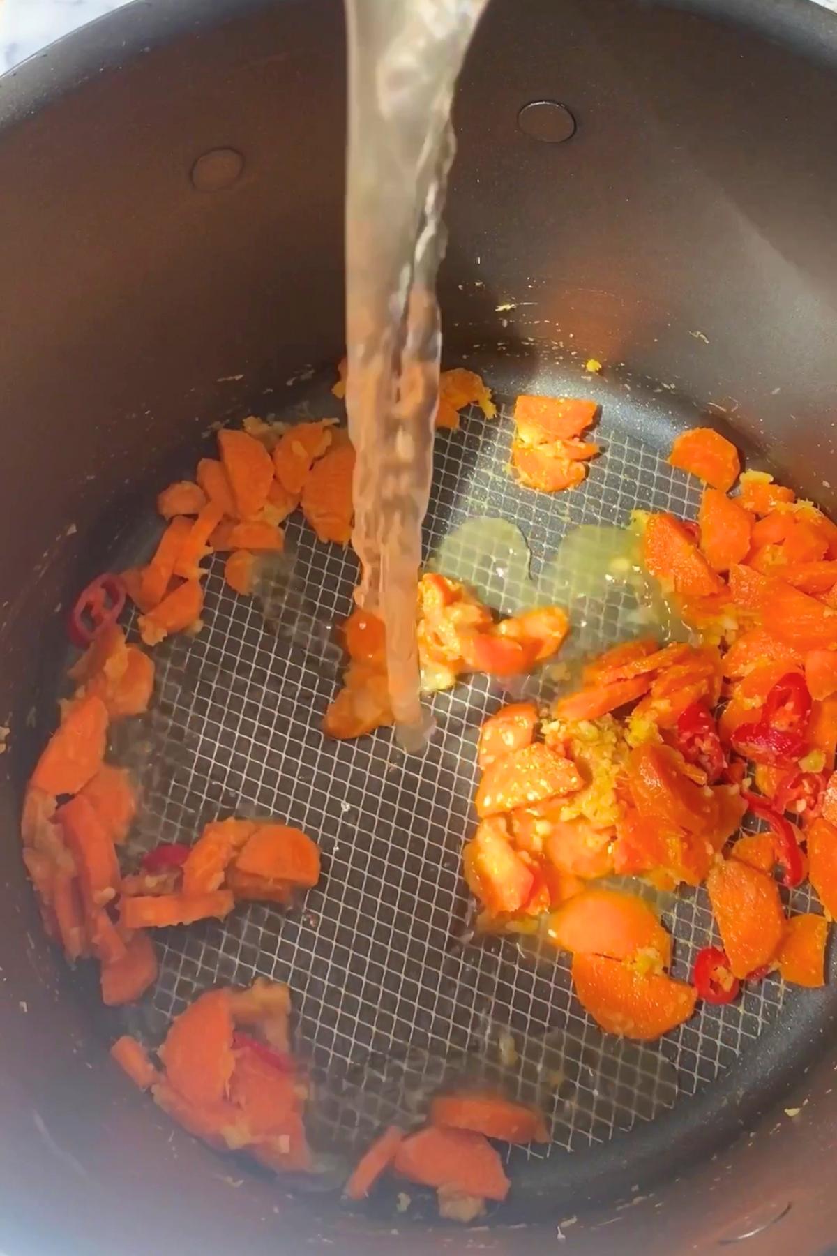 Broth pouring into a pot with sautéed carrot slices and other chopped vegetables.