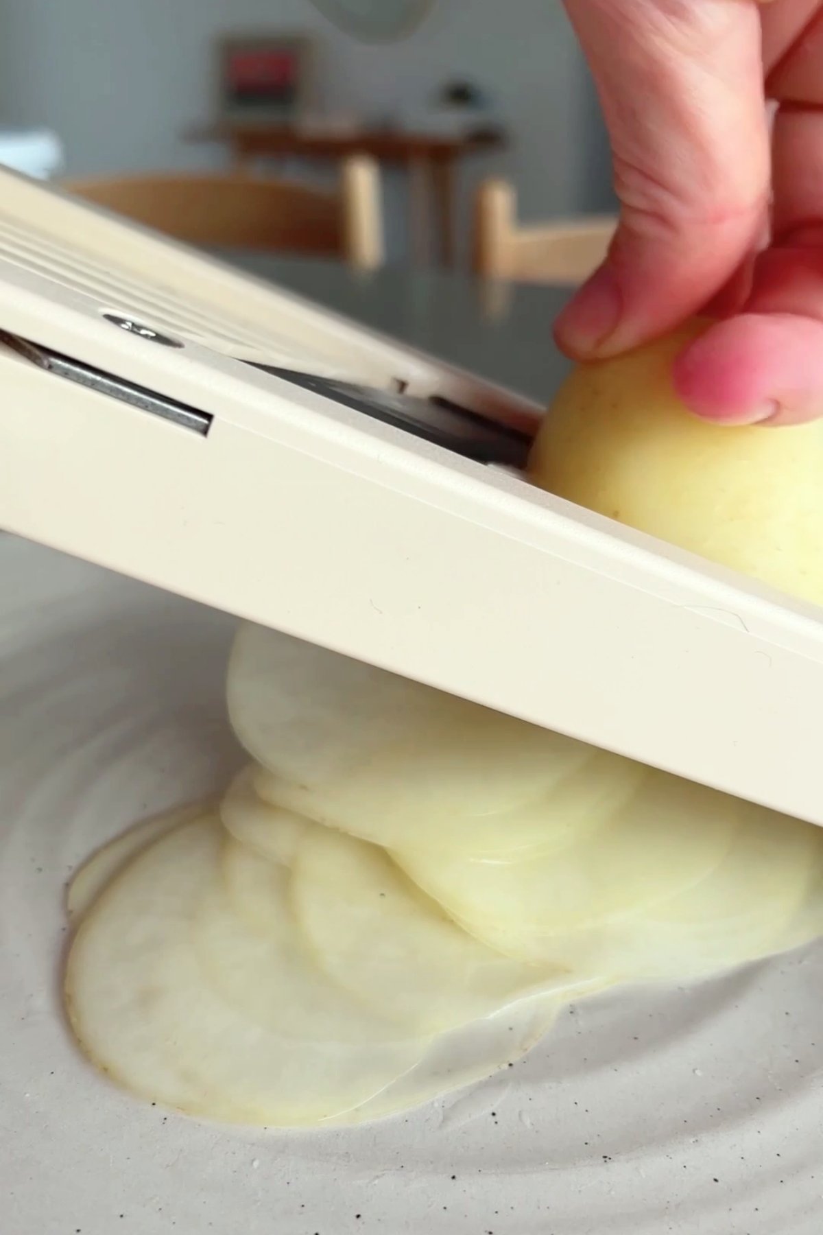 Hand pressing a potato through a mandolin, thin potato slices fanning out on a plate.