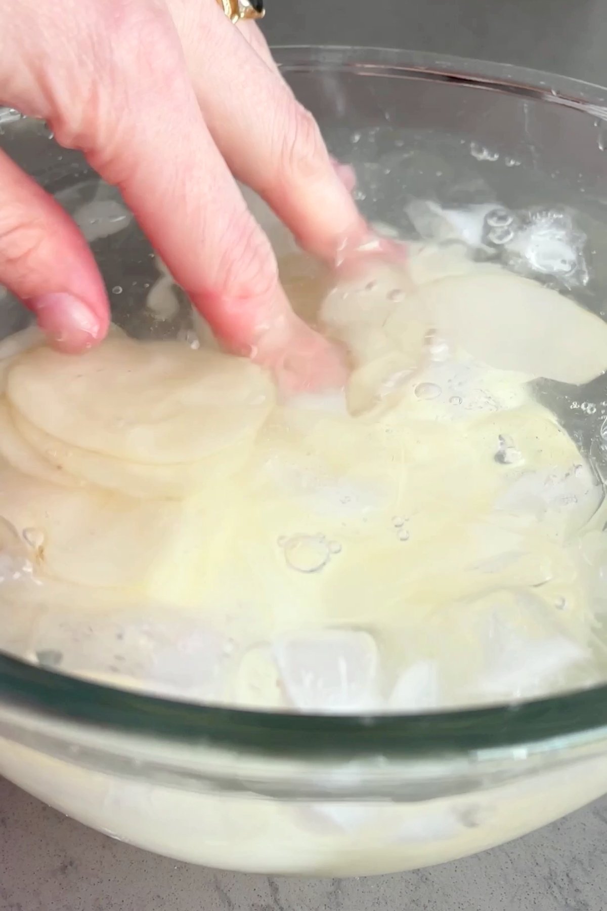 Hand adding thin potato slices in a glass bowl filled with ice water.