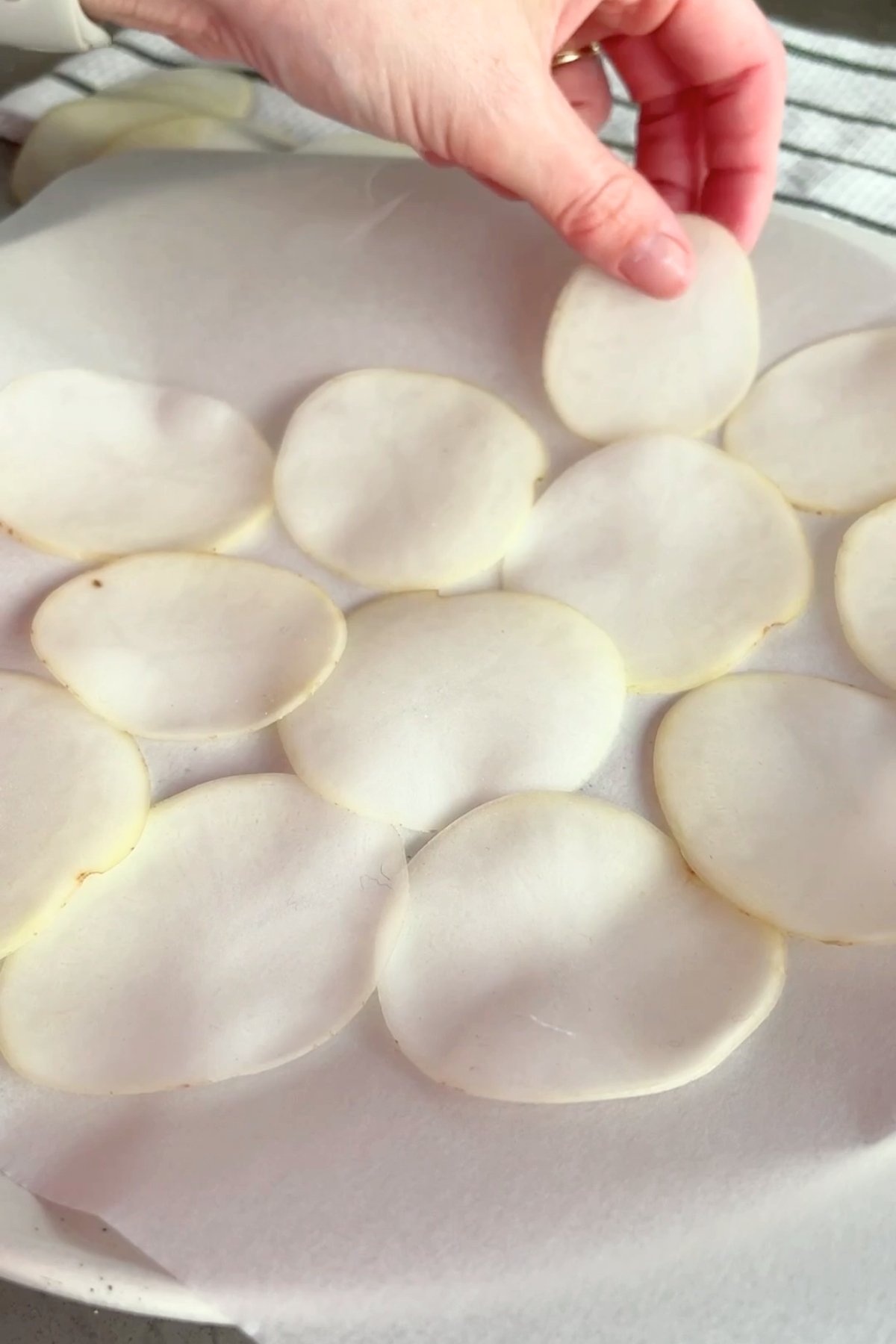 Hand arranging thin potato slices in a single layer on parchment paper.
