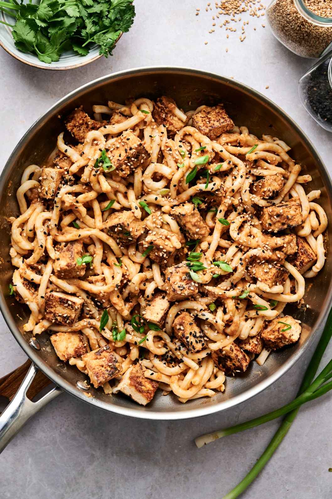 Skillet of noodles and crispy cubes coated in sauce, with scattered greens and seeds alongside.