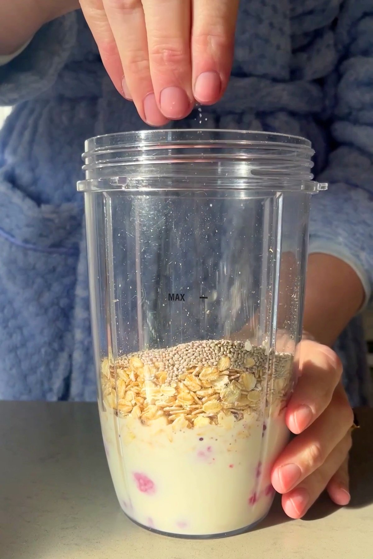 A hand sprinkles salt into a blender cup filled with oats, chia seeds, and milk with visible raspberries at the bottom.