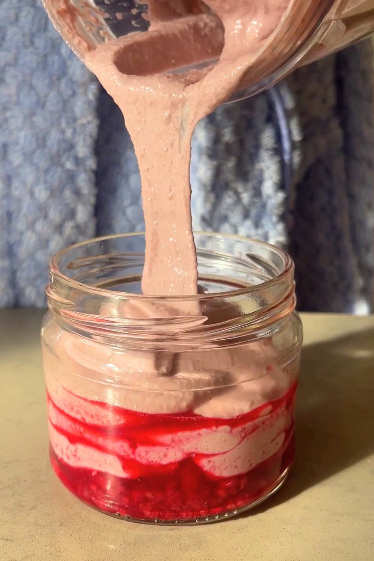 Thick pink oat mixture is being poured into a jar layered with vibrant red raspberry compote.
