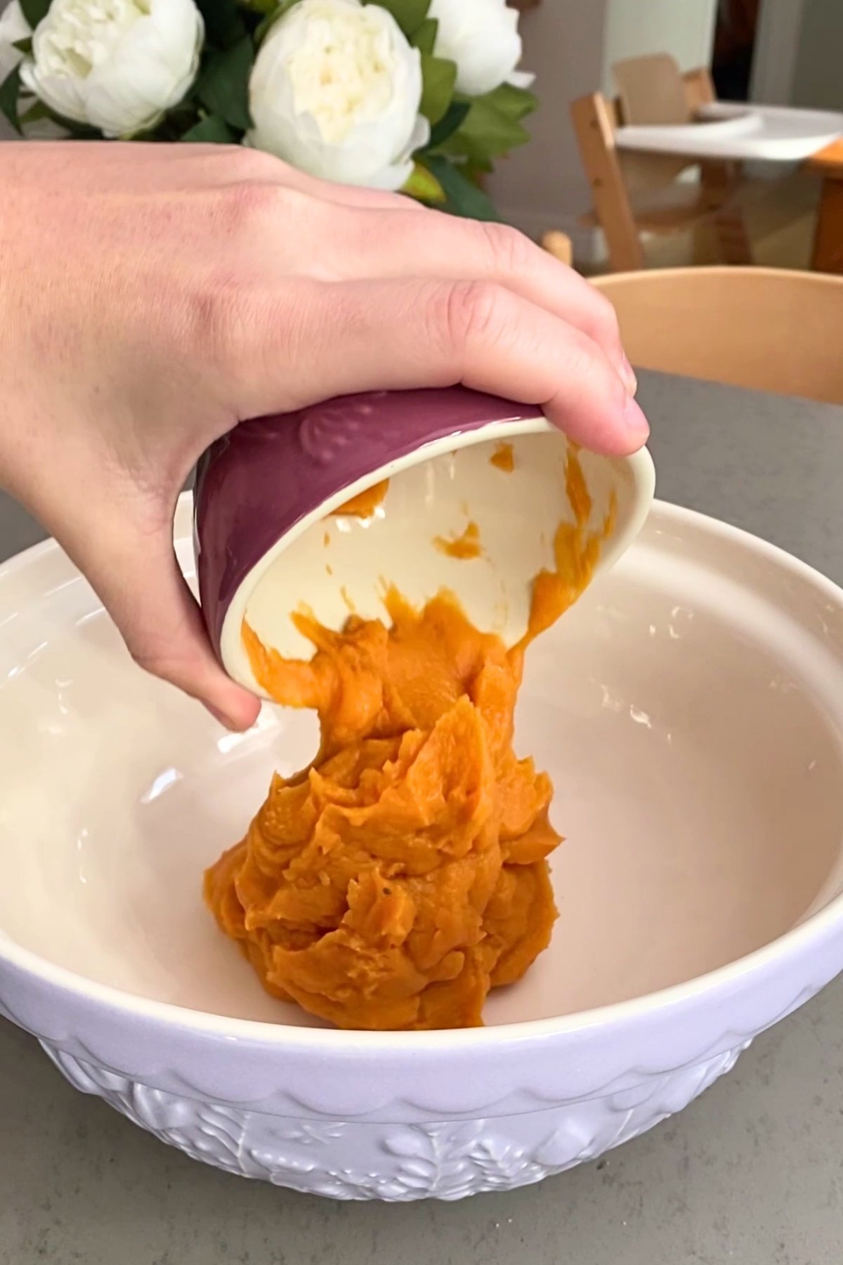 A hand pours smooth orange pumpkin puree from a small red bowl into a large white mixing bowl.