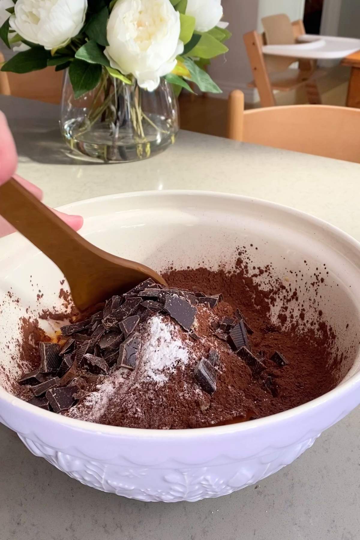 A hand mixes chocolate chunks, cocoa powder, and flour in a white bowl with a wooden spoon.