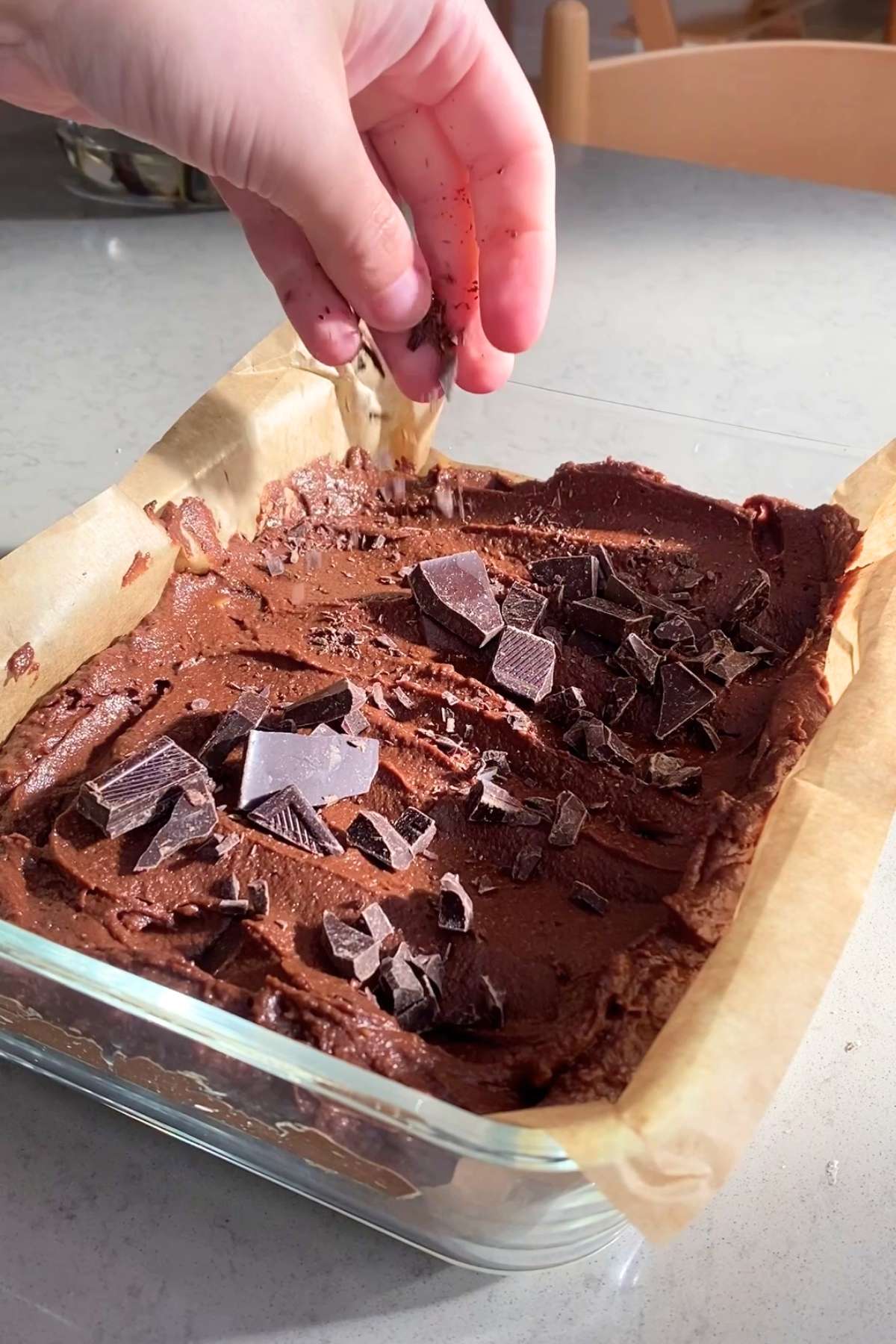 A hand sprinkles chocolate chunks onto a brown batter in a parchment-lined glass dish.