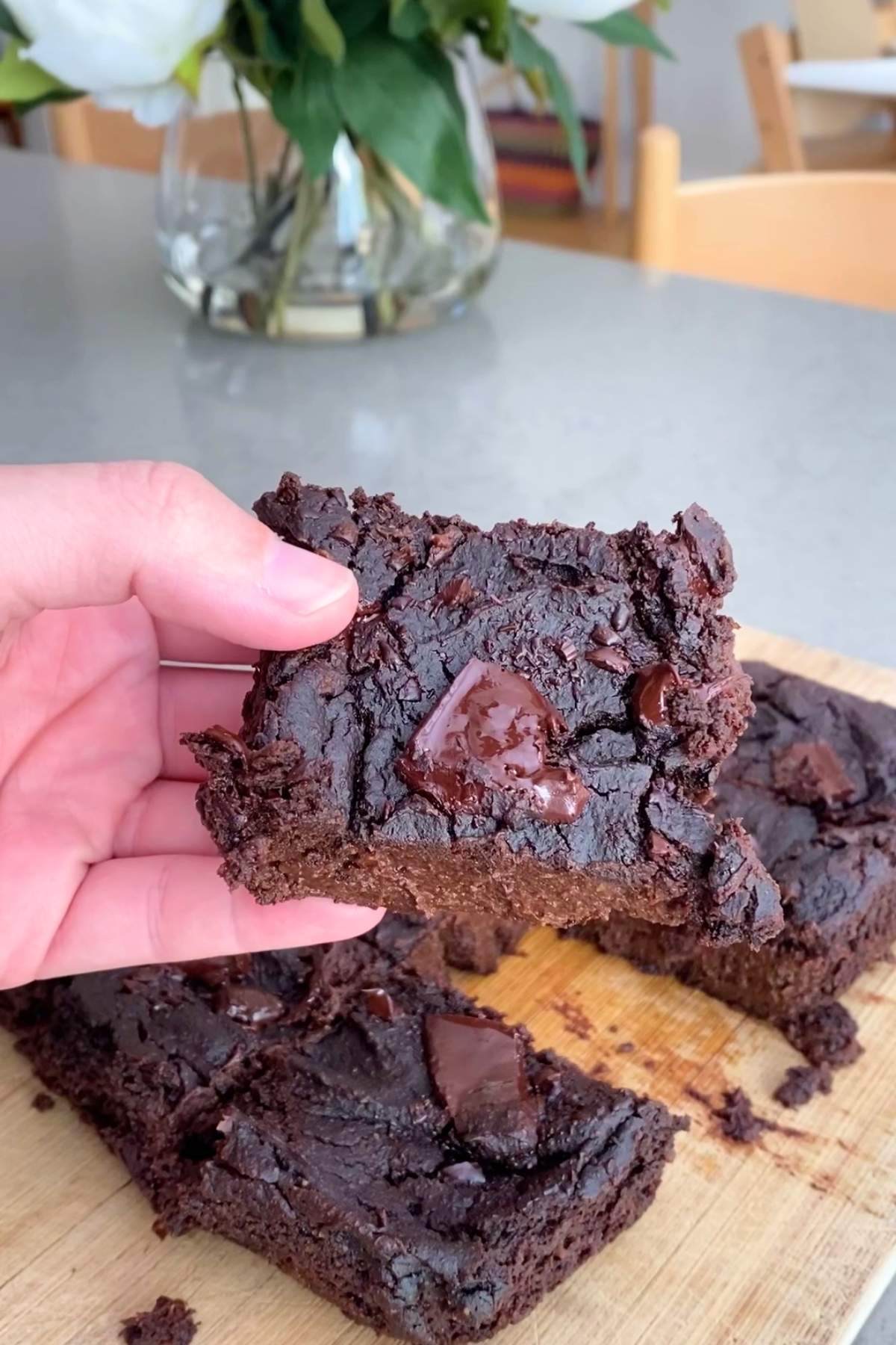 A hand holds a rich, fudgy chocolate brownie with large chocolate chunks on a wooden board.