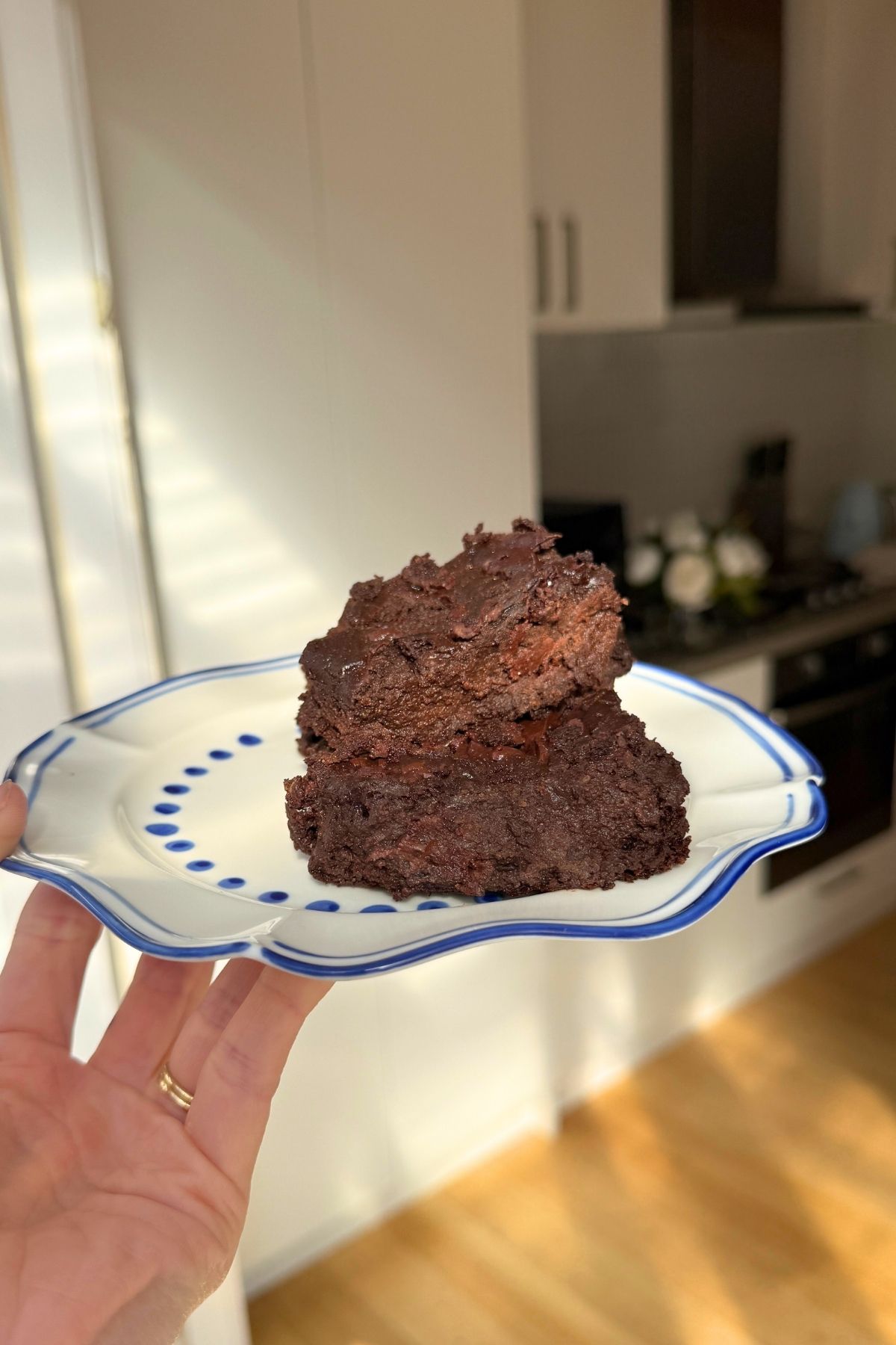 A hand holds a white plate with blue trim, featuring two rich, fudgy brownies.