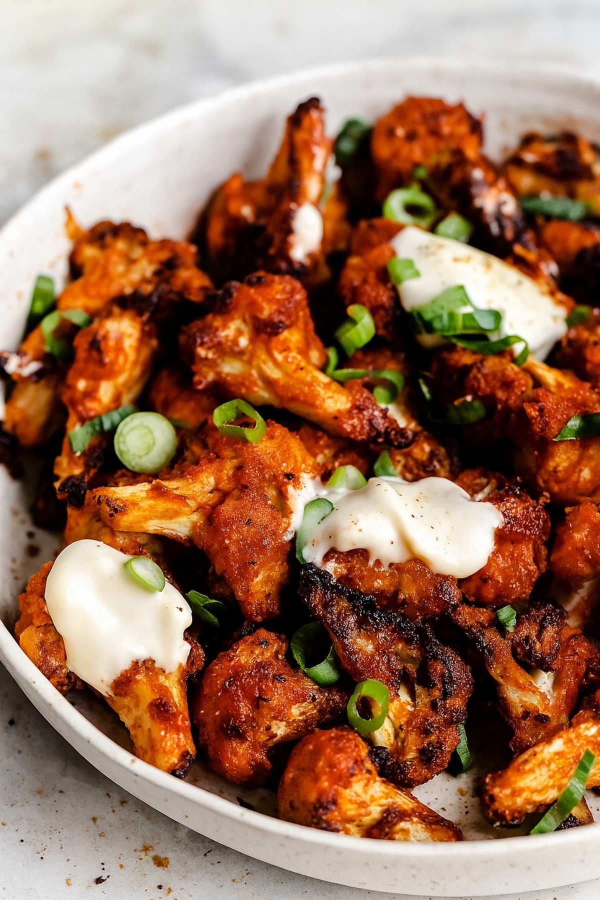 Cauliflower florets coated in bright orange sauce, garnished with chopped spring onion and dollops of creamy aioli in a bowl.