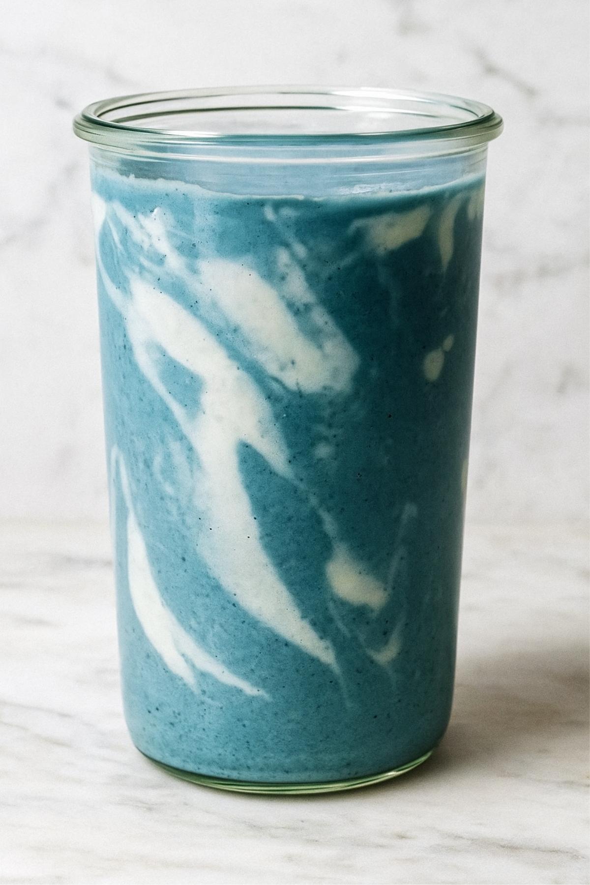 Blue smoothie with white yogurt swirls served in a tall, clear glass jar on a marble surface.