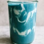 Swirled blue and white smoothie in a clear glass jar on a marble surface.