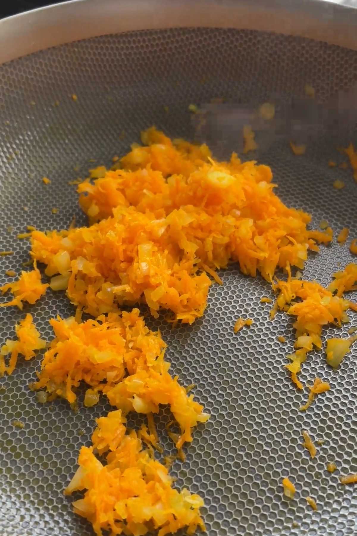 Grated carrot and onion sautรฉing in a saucepan.