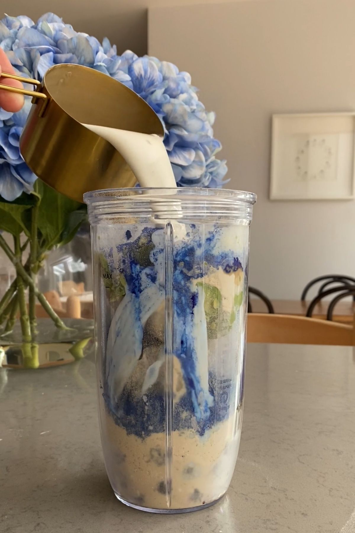 Soy milk being poured from a gold measuring cup into a blender cup filled with smoothie ingredients, including visible blue spirulina streaks.