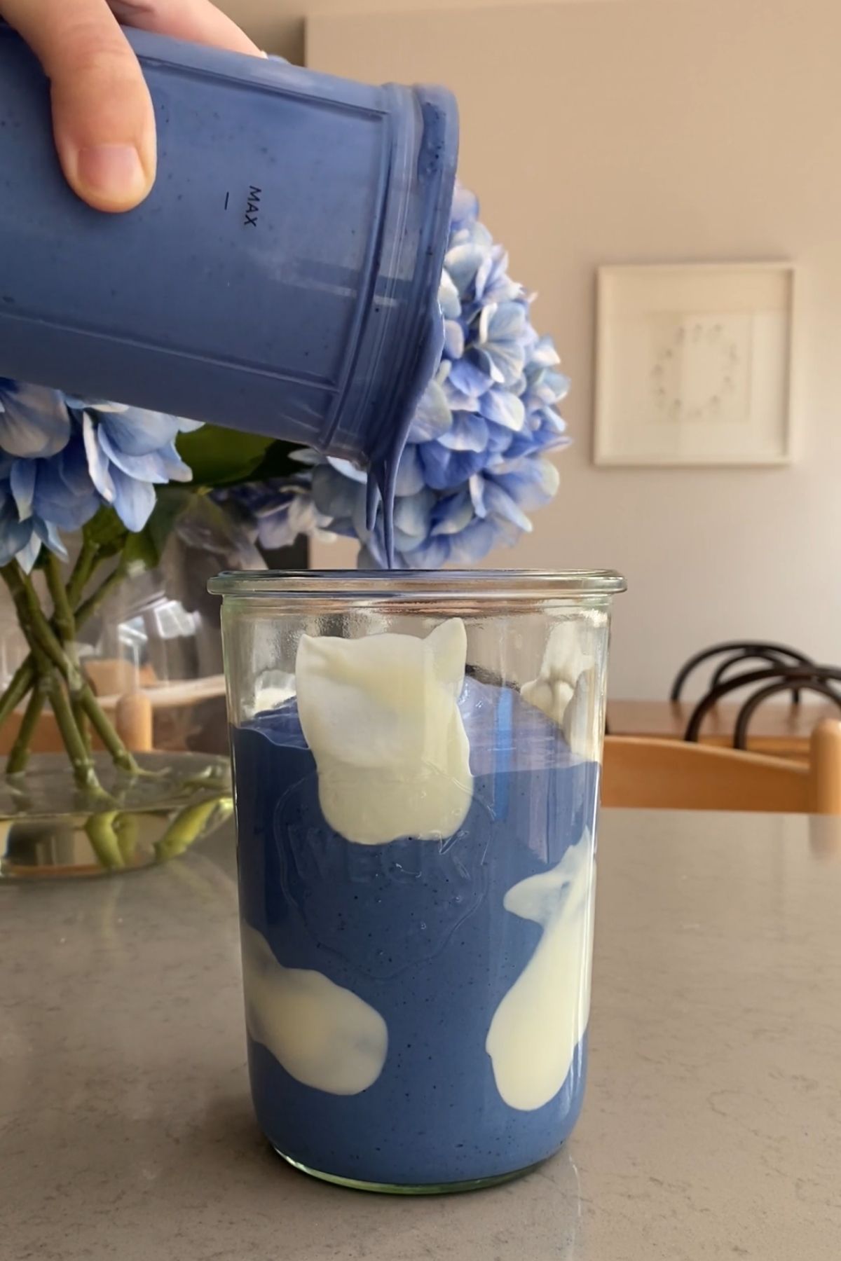 Thick blue smoothie being poured into a clear glass jar lined with white yogurt swirls, set on a kitchen counter with hydrangeas in the background.