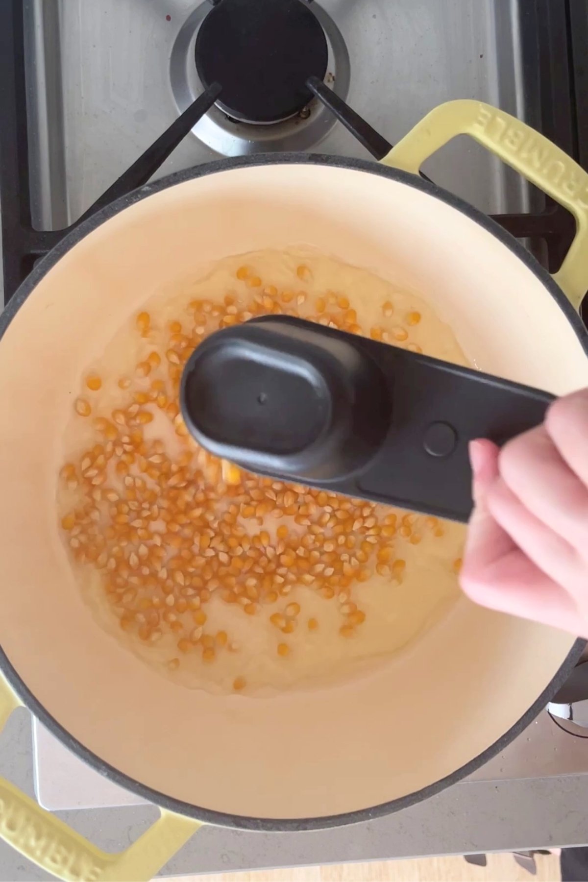 Yellow popcorn kernels heating in oil in a cream-colored pot on a stovetop, with a hand holding a measuring spoon above.