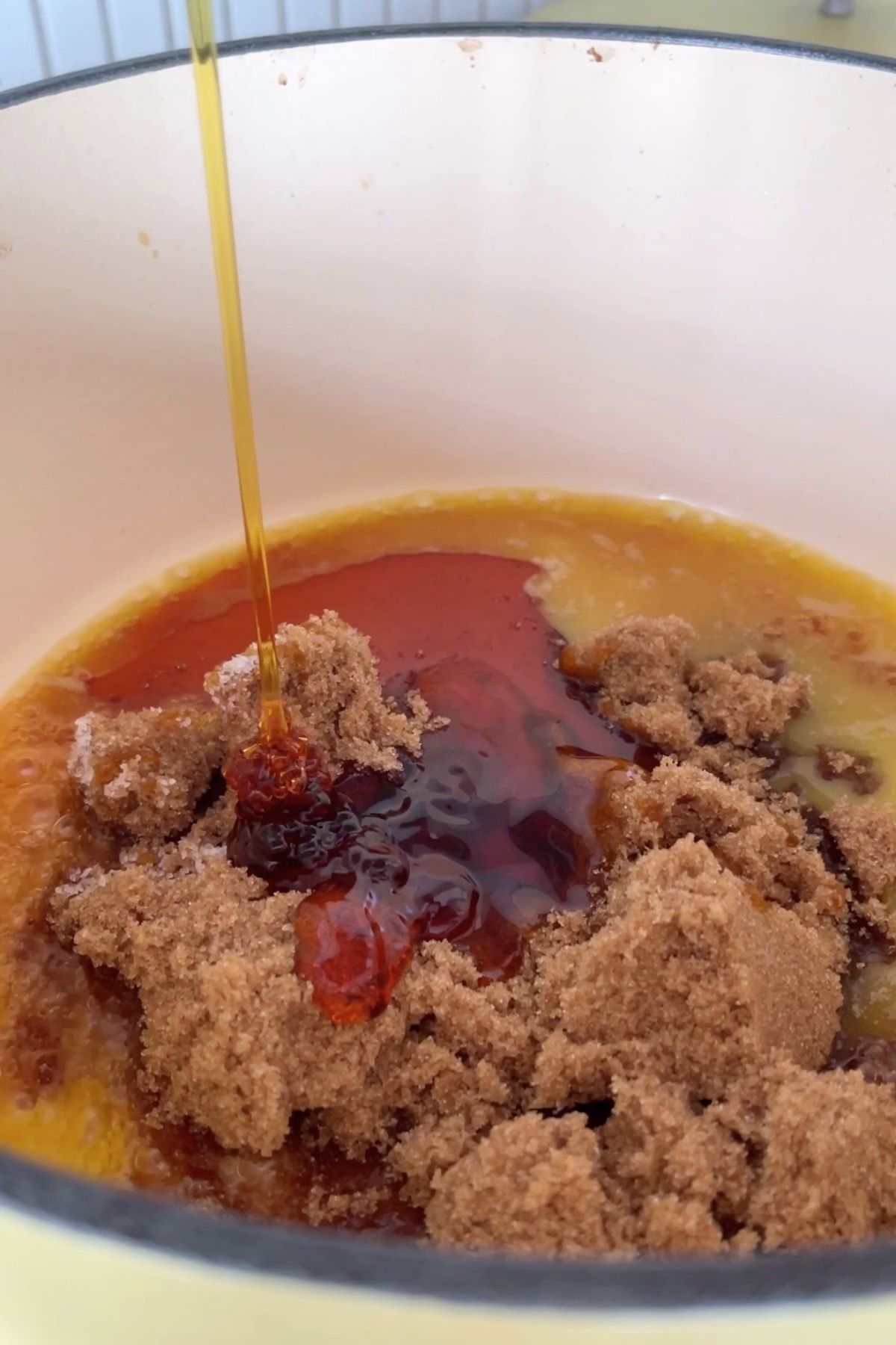Brown sugar and syrup being poured into melted butter in a cream-colored pot, forming a rich caramel mixture.