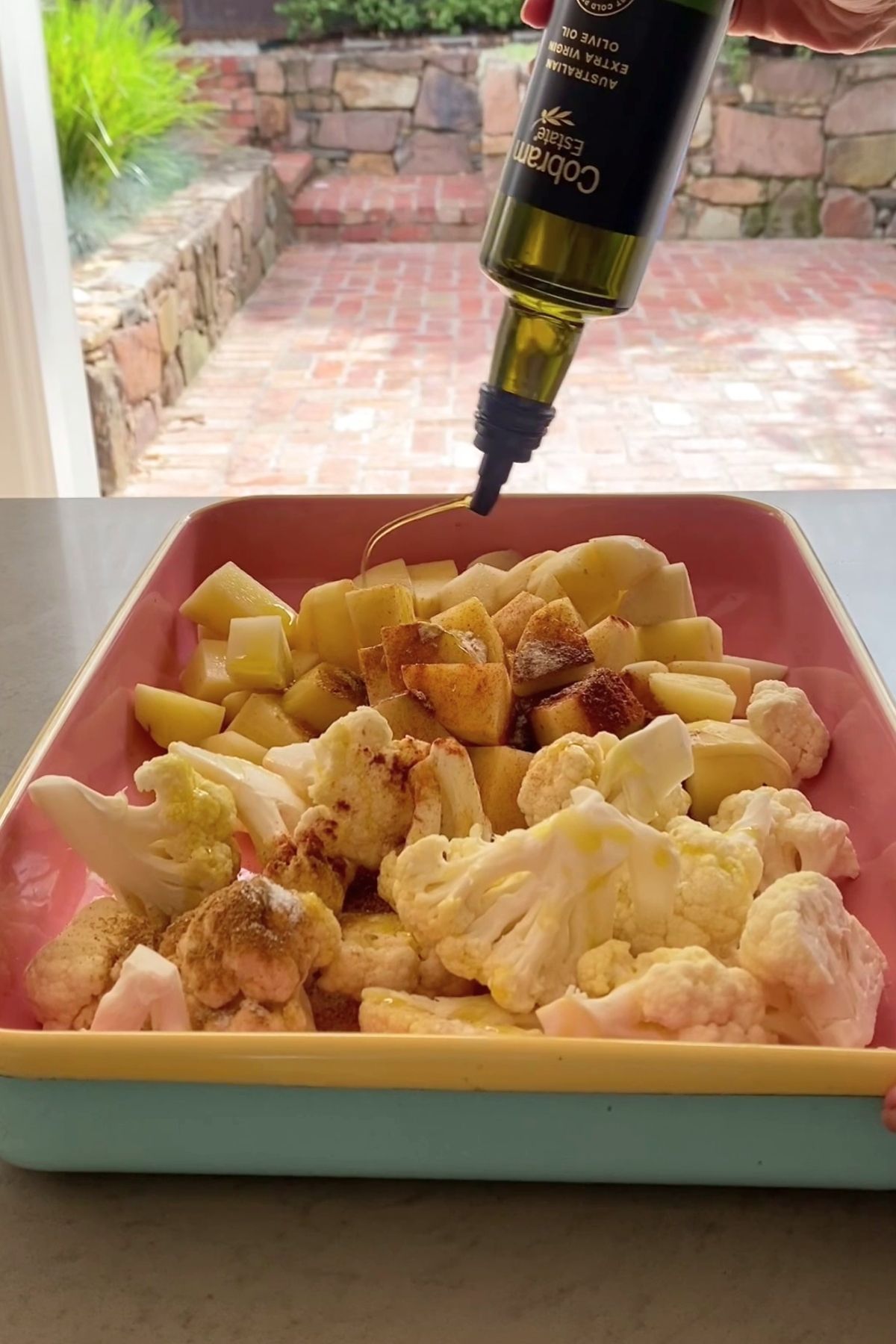 Olive oil being drizzled over seasoned cauliflower florets and cubed potatoes in a pastel roasting dish.