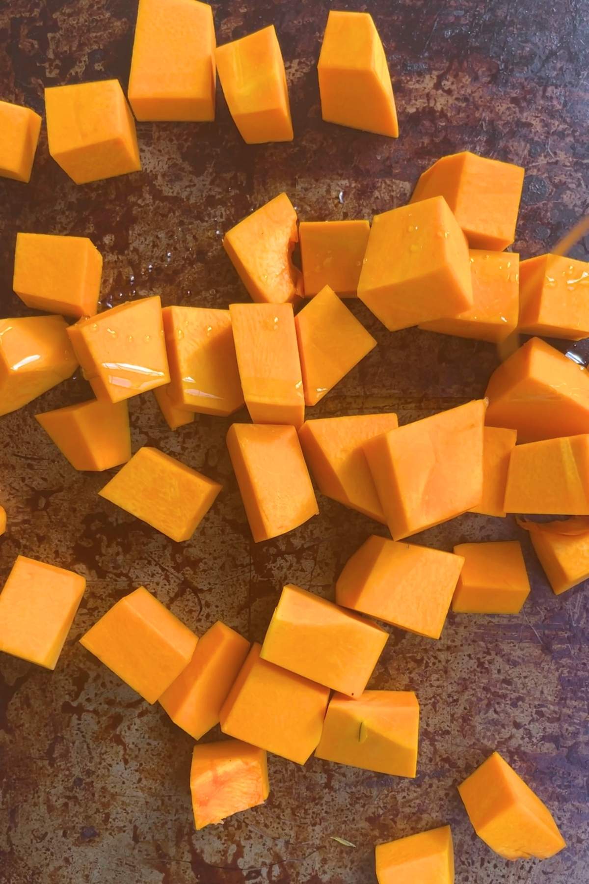 Golden cubes of raw pumpkin scattered on a well-used, dark metal baking tray.