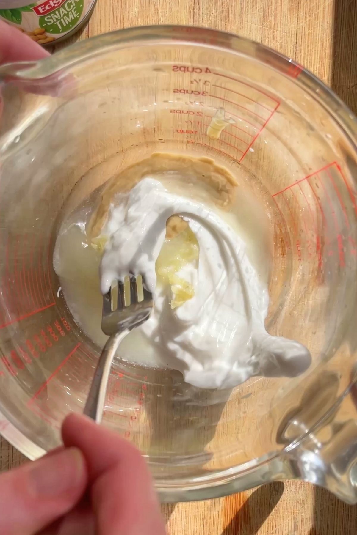 Thick white yogurt and a dollop of tahini being stirred with a fork in a glass measuring jug.