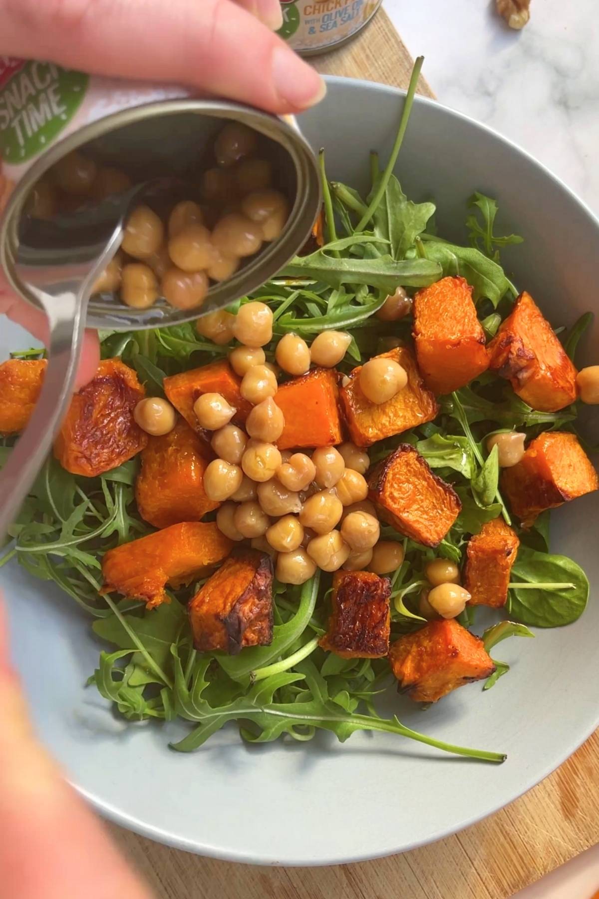 Canned chickpeas being spooned over roasted pumpkin and fresh arugula in a light blue bowl.