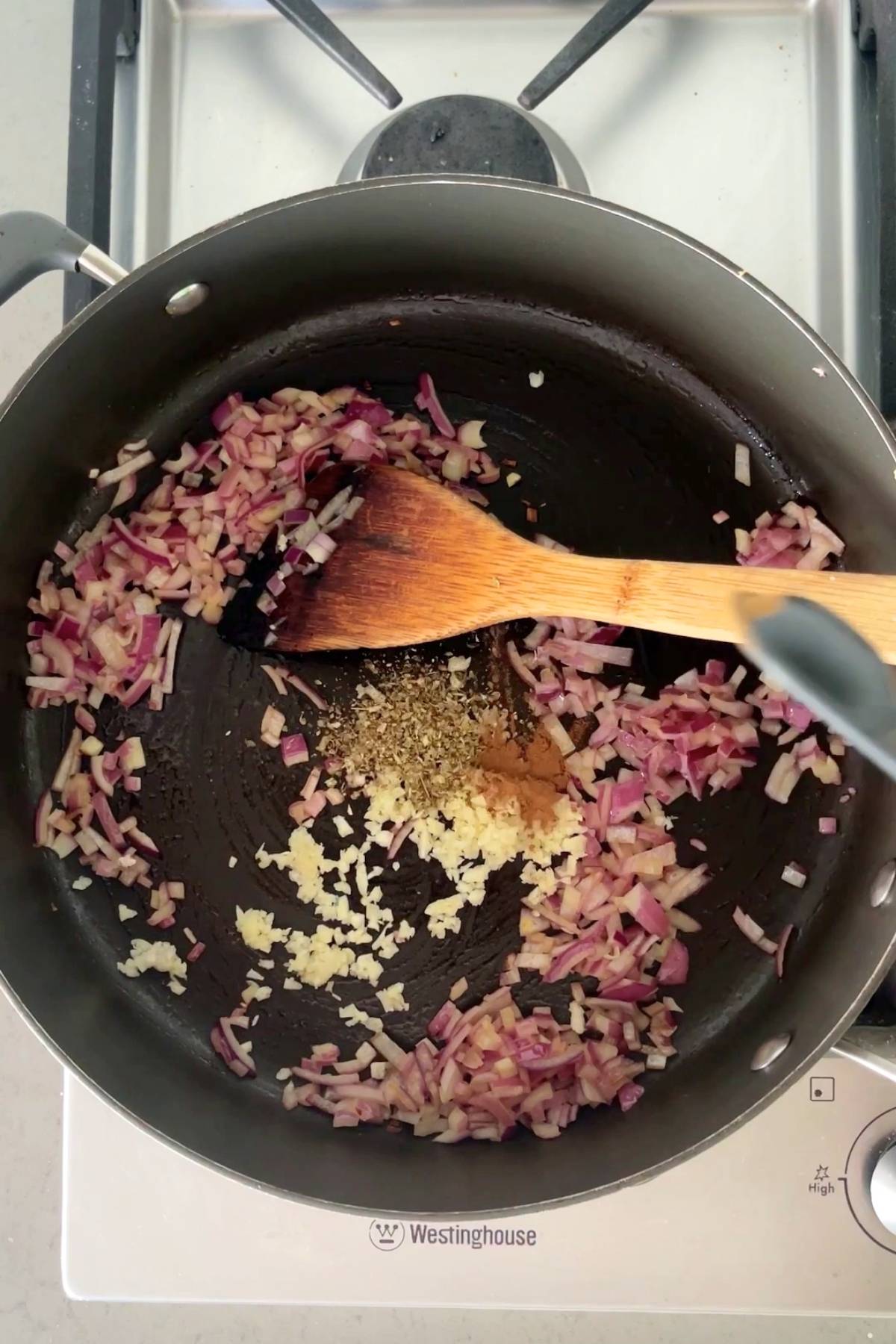 Sautéed red onion, minced garlic, and ground spices cooking in a black pan with a wooden spoon.