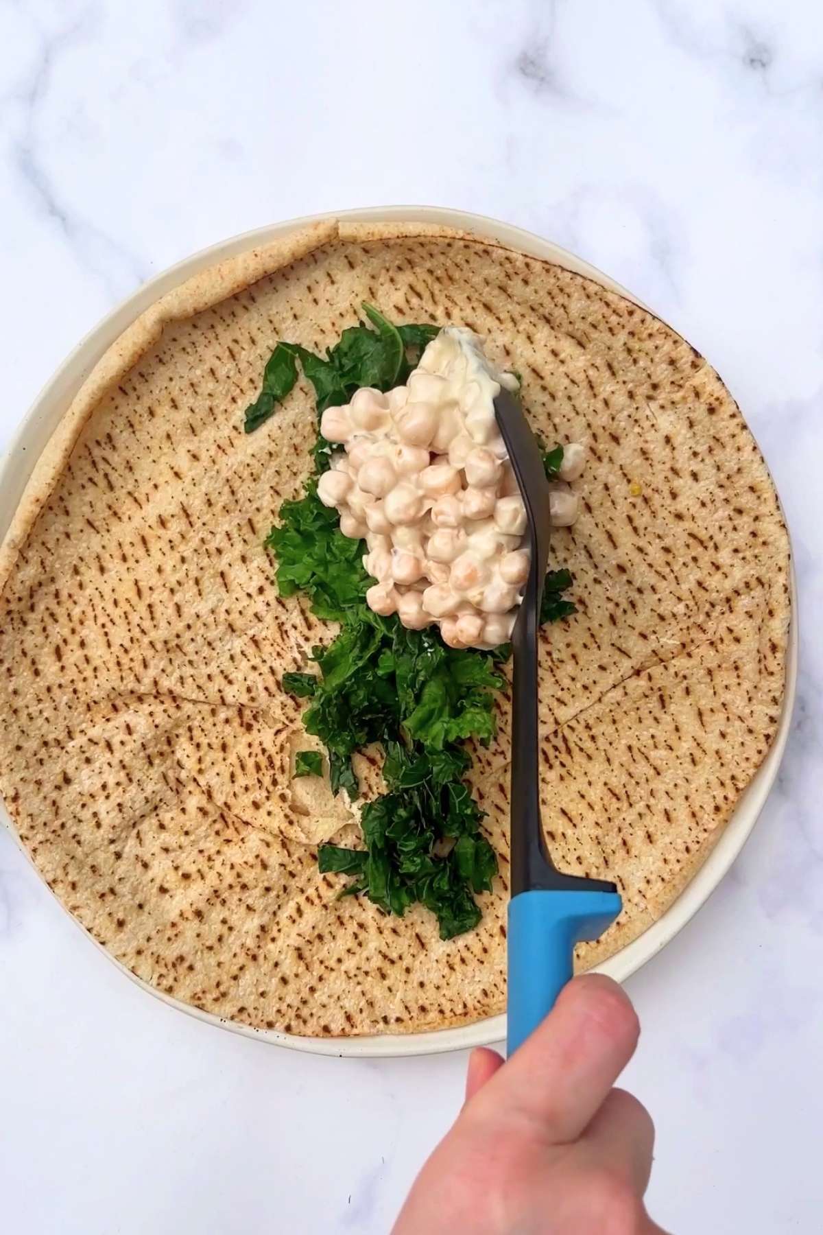 Creamy chickpeas and chopped greens being spooned onto grilled flatbread on a white plate.